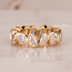 May include: A gold ring featuring a series of clear, sparkling gemstones in various shapes, including round, pear, and emerald cuts. The ring is set in a polished gold band, creating a continuous, elegant design. The ring is displayed on a light-colored surface.
