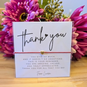 Pode incluir: Um cartão branco com as palavras "thank you" em escrita preta, uma corda vermelha e um pequeno pingente de coração dourado. O cartão inclui uma mensagem de apreço. O fundo apresenta flores cor-de-rosa.