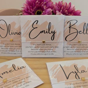 Puede incluir: Pulseras con los nombres Olivia, Emily, Bella, Amelia y Lyla en tarjetas con un diseño de pincelada color rubor. Cada tarjeta tiene un dije de corazón dorado y una cita. Las pulseras están sobre una superficie de madera.