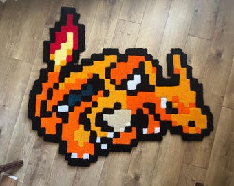 Könnte beinhalten: Ein handgefertigter, pixeliger Teppich mit dem Pokémon Glurak. Der Teppich ist hauptsächlich orange, mit schwarzen, weißen, gelben und roten Akzenten. Der Teppich liegt auf einem Holzboden.