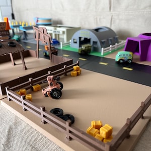 Puede incluir: Una escena de ciudad de juguete en miniatura con una valla marrón, un tractor y varios vehículos de juguete. La escena incluye una carretera, edificios y pequeños bloques amarillos. La paleta de colores es apagada, con tonos marrones, beiges y grises.