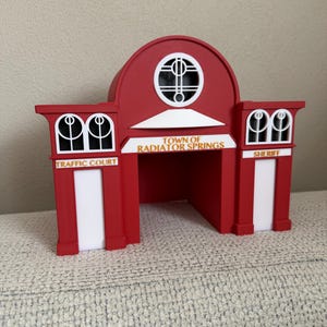 以下が含まれることがあります： 「Town of Radiator Springs」の文字が入った、赤色の玩具の建物。白いアクセントがあり、「Traffic Court」と「Sheriff」の標識があります。入り口の上には円形の窓があります。