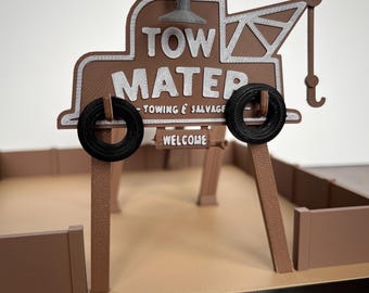 Puede incluir: Un modelo de cartón marrón y blanco de una grúa de remolque con las palabras "Tow Mater" y "Towing & Salvage" en el lateral. El modelo está sobre una base marrón con postes de madera marrón.
