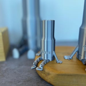 Puede incluir: Tres figuras de personas impresas en 3D de color plateado sentadas sobre una superficie de madera. Las figuras tienen piernas y brazos y están etiquetadas con las palabras "0mm", "1mm" y "2mm".