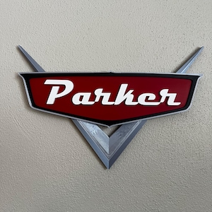 Puede incluir: Un letrero rojo y negro con el nombre "Parker" en letras blancas. El letrero tiene una base plateada estilizada en forma de V con dos alas que apuntan hacia arriba. El letrero está montado en una pared texturizada de color claro.