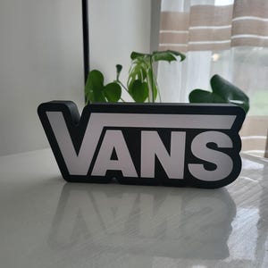 Peut inclure: Enseigne lumineuse avec le logo Vans en noir et blanc. L'enseigne est en plastique et a une bordure noire. Le logo Vans est blanc et est éclairé.