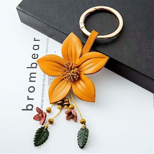 May include: A leather flower keychain in shades of orange, with a gold-colored key ring. The flower has detailed petals and a textured center. Small red flowers and green leaves dangle below. The word "brombear" is visible.