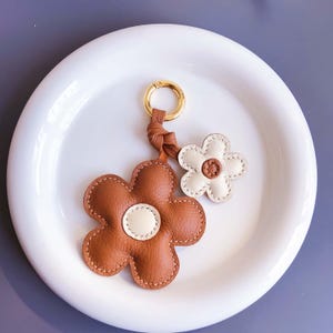 May include: A leather flower keyring with a gold-coloured ring. The keyring features two flower charms, one brown and one white, with cream-coloured centres. The flowers are stitched with a contrasting thread.