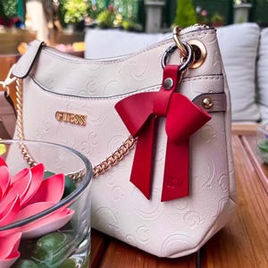 Könnte beinhalten: Cremefarbene Guess-Handtasche mit goldener Kette und geprägtem Logo. Ein leuchtend roter, schleifenförmiger Lederanhänger hängt am Griff. Die Tasche steht auf einer Holzoberfläche.
