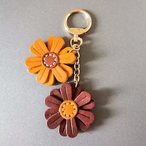 May include: A leather keychain featuring two flower-shaped charms. One charm is orange, the other is brown, both with yellow centers and gold-colored hardware. The keychain is attached to a gold-colored clasp and chain.