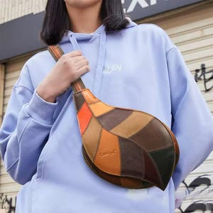Puede incluir: Un bolso bandolera en forma de lágrima confeccionado con un mosaico de piezas de cuero en varios tonos de marrón, beige y naranja. El bolso cuenta con una correa de cuero a juego y costuras visibles a lo largo de los bordes de cada panel.