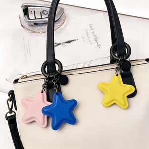 May include: A cream-colored handbag with black handles and a silver zipper. Attached are three star-shaped keychains in pink, blue, and yellow. A car key and a book are in the background.
