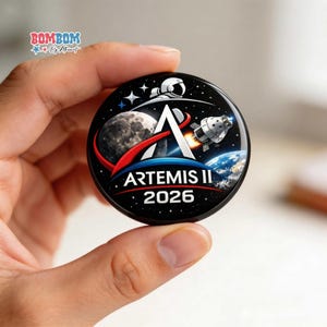 May include: A round, black Artemis II 2026 button featuring a space-themed design. The design includes an astronaut, a rocket, Earth, the moon, and the Artemis II logo in white and red.