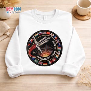 Puede incluir: Sudadera blanca de cuello redondo con un parche circular con un diseño de cohete, el texto "PROJECT HAIL MARY" y el nombre "MARY". El parche está rodeado de varias banderas nacionales. La sudadera tiene mangas largas.