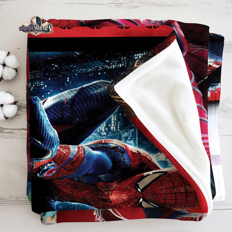 Puede incluir: Una manta plegada con una imagen vibrante de Spider-Man con su ic&oacute;nico traje rojo y azul, sobre un fondo de paisaje urbano. La manta tiene un borde rojo y un interior blanco.
