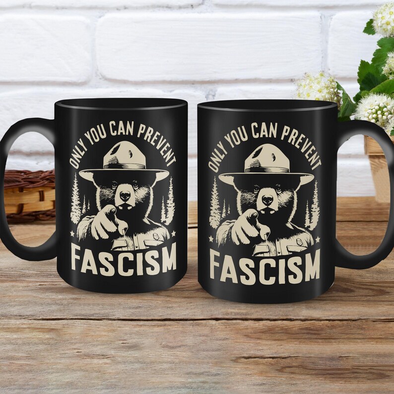 May include: Two black coffee mugs with a graphic of a bear wearing a ranger hat and pointing at the viewer. The text on the mugs reads "Only you can prevent fascism".
