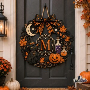 May include: A circular Halloween wreath featuring a black cat wearing a witch hat, a central letter M, a pumpkin, two potion bottles, a crescent moon, and stars. The wreath is decorated with orange autumn leaves and a large black and orange bow.