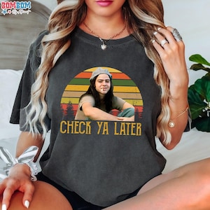 May include: Dark gray t-shirt featuring a retro-style graphic of a person with a cigarette, set against a sunset backdrop. The text "CHECK YA LATER" is printed below the image. The shirt has short sleeves.