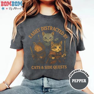 May include: Dark gray t-shirt with a graphic featuring three cartoon cats in medieval attire, holding swords. The text reads "EASILY DISTRACTED BY CATS & SIDE QUESTS" in orange. The shirt also has the words "COMFORT COLORS" and "PEPPER" in a circle.
