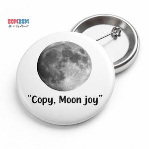 May include: White button pin with a full moon graphic and the text "Copy, Moon joy". The pin has a silver metal back with a safety pin closure. The pin is a round shape.