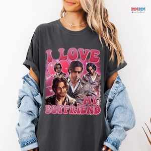 May include: Dark gray t-shirt with a pink graphic that reads "I LOVE MY BOYFRIEND." The graphic features a heart and multiple images of a man. The shirt has short sleeves and a relaxed fit.