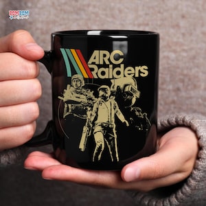 May include: Black ceramic mug featuring the text "ARC Raiders" with a retro color scheme and illustrations of soldiers. The mug has a black handle and is held by a person.