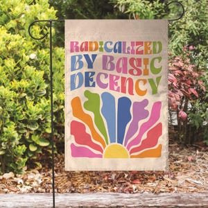 May include: A beige garden flag with the text "RADICALIZED BY BASIC DECENCY" in colorful, retro-style lettering. A stylized sunburst design in shades of orange, pink, green, and blue is below the text. The flag is displayed on a black metal stand.