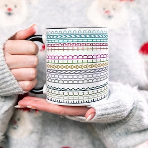 May include: A white ceramic mug with a black handle and rim, featuring a colorful, patterned design of various stitched lines. The mug is held by a person wearing a gray sweater.