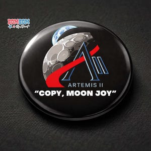 May include: A black button featuring the Artemis II logo with a stylized moon and Earth graphic. The design includes the text "ARTEMIS II" and the phrase "COPY, MOON JOY" in white lettering. A red stripe accents the logo.