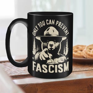 May include: Black coffee mug with a white graphic of a bear wearing a ranger hat and pointing at the viewer. The text "Only You Can Prevent Fascism" is printed on the mug.