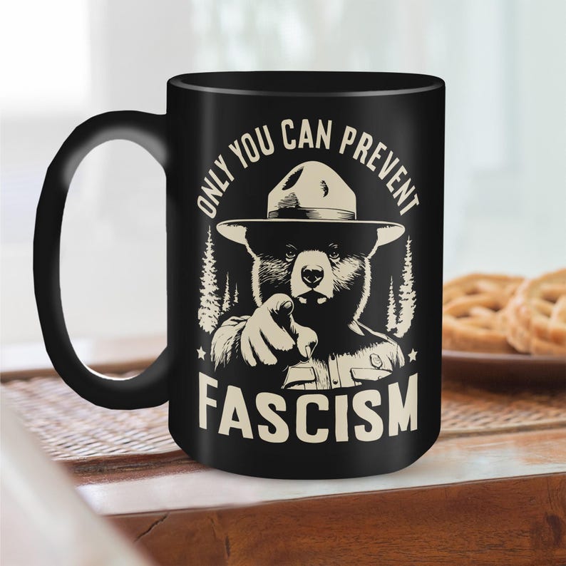 May include: Black coffee mug with a white graphic of a bear wearing a ranger hat and pointing at the viewer. The text "Only You Can Prevent Fascism" is printed on the mug.