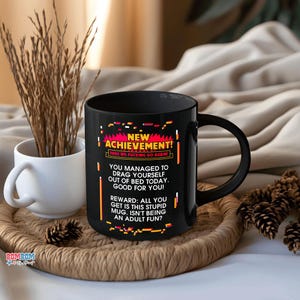 May include: Black ceramic mug with the text "NEW ACHIEVEMENT!" and a humorous message about getting out of bed. The mug is on a woven tray with a white mug and decorative pinecones.