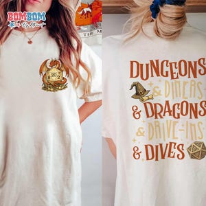 May include: Cream-colored t-shirt featuring a dragon and a 20-sided die graphic. The back of the shirt displays the text "Dungeons & Dragons & Drive-Ins & Dives" with a wizard hat and die graphic. A fun, casual tee.
