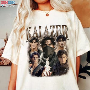 May include: Cream-colored t-shirt featuring the word "KALAZER" in a brown, metallic font. The shirt displays a graphic of five male figures with dark hair, some wearing scarves or cloaks. The design has a fantasy theme.