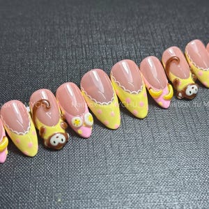 May include: A set of ten press-on nails featuring a French manicure style with a nude base and yellow tips adorned with pink polka dots. Some nails include 3D embellishments of brown monkeys and yellow bananas, with the text Nailbox by MYCHU.