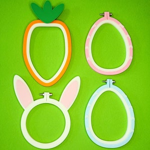 May include: Four embroidery hoops in different shapes and colors. A carrot shaped hoop, a pink oval hoop, a blue oval hoop, and a white bunny shaped hoop.