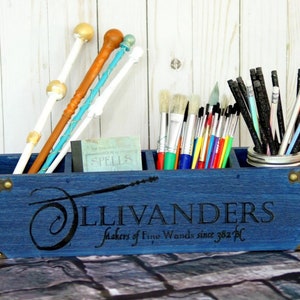 Peut inclure: Organisateur en bois bleu avec trois compartiments, étiqueté "Ollivanders Makers of Fine Wands since 382 BC". Il contient des pinceaux, des crayons et quatre baguettes.