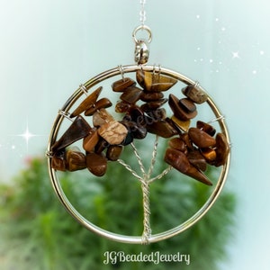 May include: A silver-toned metal tree of life pendant. The tree's branches and leaves are made of brown and tan tiger eye stones. The pendant is suspended from a delicate silver chain. The text "JG Beaded Jewelry" is at the bottom.