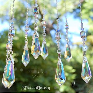 May include: Multiple strands of iridescent crystal pendants suspended from silver chains. Each pendant has a unique shape and a faceted surface that reflects light. The background is a blurred view of green leaves. The text "JG Beaded Jewelry" is at the bottom.