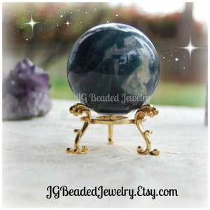 May include: A dark green and white crystal ball sits on a gold-colored decorative stand. The stand has ornate, curved legs. The text "JG Beaded Jewelry.Etsy.com" is at the bottom of the image. The background is blurred.
