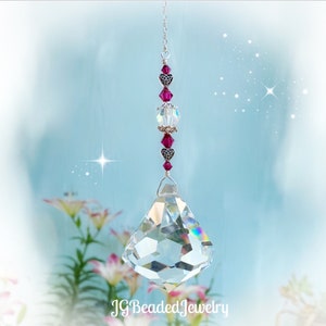May include: A clear crystal prism suncatcher with a silver chain and decorative beads. The beads are a mix of clear, pink, and silver, with heart-shaped accents. The suncatcher hangs in front of a soft blue background with flowers.