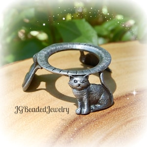 May include: A silver-colored metal ring holder with a cat figurine. The ring holder has a circular top supported by a seated cat and two curved supports. The text "JG Beaded Jewelry" is visible at the bottom.