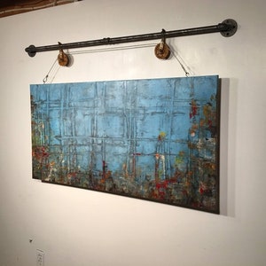 This painting has a grid pattern , a blue backround and multiple colors along the bottom in of red, green, mustard, orange. It is finished with a dark wash and hangs from a black iron pole that has a metal rope and rusted pulleys.Its highly textured.