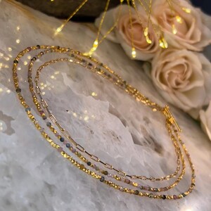 Può includere: Una collana dorata a più strati con tre fili di piccole perle e una delicata catena d'oro. Le perle sono un mix di rosa, verde e nero.