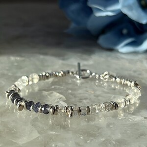 Könnte beinhalten: Ein filigranes Silberarmband mit Knebelverschluss. Es ist mit einer Mischung aus klaren und dunkelblauen Edelsteinen verziert, die von kleinen Silberperlen unterbrochen werden. Die Edelsteine haben unterschiedliche Größen und Formen.