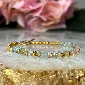 Könnte beinhalten: Ein zartes Armband mit kleinen, unregelmäßig geformten Edelsteinen in Blau-, Grün- und Rosatönen. Die Edelsteine sind mit kleinen, goldfarbenen Perlen durchsetzt und durch einen goldenen Verschluss verbunden.
