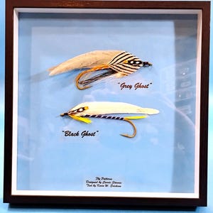 May include: Framed fly fishing lures, "Grey Ghost" and "Black Ghost", with detailed feather and thread work. The "Grey Ghost" lure has a tan wing and striped head. The "Black Ghost" lure is white with yellow accents. The frame is dark brown.