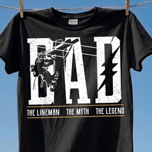 Op de afbeelding: Zwart T-shirt met het woord "DAD" in grote, verweerde witte letters. De letter "A" heeft een lineman-afbeelding. Daaronder staat de tekst "THE LINEMAN THE MYTH THE LEGEND" in goud.