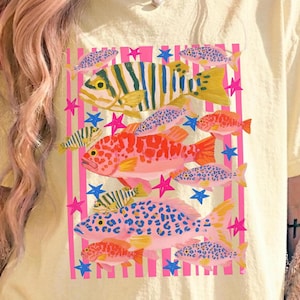 Puede incluir: Una camiseta amarillo claro con un diseño de peces coloridos. El gráfico incluye varios peces en tonos rosa, naranja, azul y amarillo, sobre un fondo de rayas rosas y estrellas azules. Los peces tienen patrones y texturas únicos.