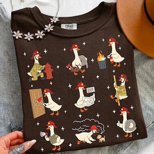 Könnte beinhalten: Ein dunkelbraunes T-Shirt mit einem wiederkehrenden Grafikmuster aus weißen Gänsen, die als Feuerwehrleute verkleidet sind. Die Gänse sind bei verschiedenen Aufgaben dargestellt, halten Äxte, Feuerwehrschläuche, Feuerlöscher und Pläne und tragen rote Helme.
